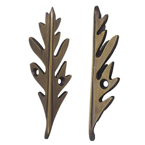 May include: Two bronze-coloured leaf-shaped decorative metal pieces. Each piece has a detailed leaf vein design with three holes for mounting. The leaves are symmetrical and have a vintage or antique appearance.