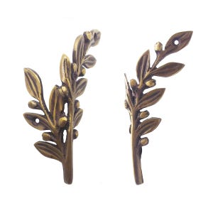 Puede incluir: Dos adornos decorativos de metal color bronce con forma de hojas. Cada pieza presenta hojas detalladas y pequeñas decoraciones en forma de brotes. Los adornos tienen agujeros para el montaje y están sobre un fondo blanco.