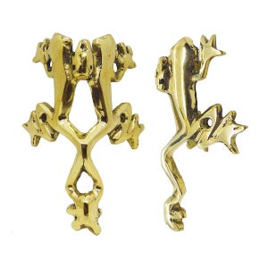 May include: Two brass frog-shaped decorative objects. One frog is upright, the other is sideways. The frogs have detailed features, including textured skin and star-shaped feet. The objects are likely used as decorative hardware or embellishments.