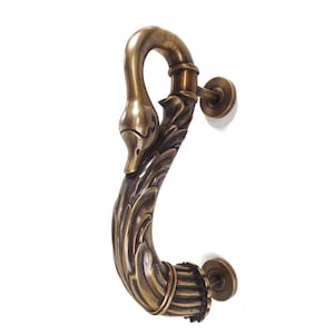May include: A bronze-colored door handle in the shape of a swan. The handle features a swan's head and neck, with a curved body adorned with leaf-like details. The handle is mounted on a white surface.