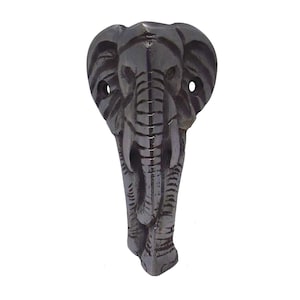 May include: A dark gray, carved wooden elephant head with tusks and detailed trunk. The elephant has two holes for mounting. The carving is a front-facing view of the elephant's head and trunk.