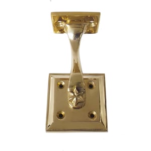 Puede incluir: Un tirador de ventana decorativo de latón. Tiene una base rectangular con cuatro orificios para tornillos, un brazo curvo y una sección superior con dos orificios. Una estrella está grabada en el brazo. Mide unos 10 cm.