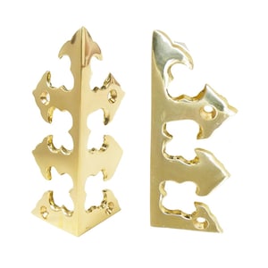May include: Two brass-coloured corner brackets with ornate, openwork designs. Each bracket has a right-angle shape with decorative cutouts and a polished finish. The brackets are designed for furniture or decorative applications.