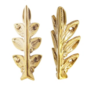 Puede incluir: Dos elementos decorativos dorados en forma de hoja. Cada elemento tiene un tallo central con varias protuberancias en forma de hoja. Las hojas tienen una superficie texturizada y pequeños agujeros para la fijación. El diseño general es simétrico y ornamentado.