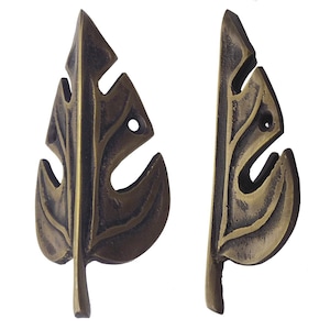 May include: A pair of bronze-coloured leaf-shaped cabinet pulls. Each pull features a detailed, textured design with a central vein and cut-out sections. The pulls are made of metal and have a vintage or antique appearance.