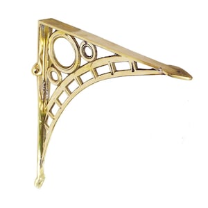 May include: A brass-colored shelf bracket with an Art Deco design. The bracket features a triangular shape with decorative circular and rectangular cutouts. The support arm extends horizontally, and the bracket has a small, decorative hook at the bottom.