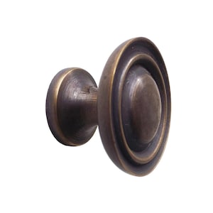 Puede incluir: Un tirador de armario de color bronce con un diseño circular. El tirador tiene una base redonda y una cara decorativa en capas. El acabado tiene un aspecto ligeramente envejecido, lo que sugiere un estilo vintage. Ideal para la restauración de muebles.