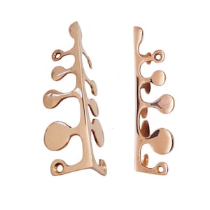 Pode incluir: Duas peças decorativas de metal em ouro rosa. Cada peça tem um design orgânico, semelhante a um galho, com elementos arredondados em forma de gota. As peças têm pequenos orifícios para fixação, sobre fundo branco.