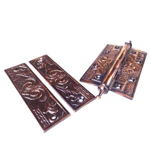 May include: Ornate bronze-colored door hinges with detailed floral and geometric patterns. The hinges are rectangular with rounded edges and include a central barrel and decorative finial. The hardware is displayed against a white background.
