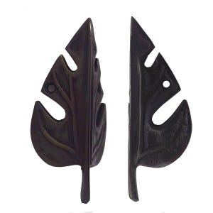 May include: Two dark bronze leaf-shaped cabinet pulls. Each pull features a detailed leaf vein design with cut-out sections. The pulls are designed to be mounted on a cabinet door or drawer, adding a decorative touch.