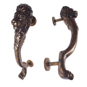 May include: Two bronze-colored door handles. Each handle is sculpted in the shape of a lion, with the lion's head at the top and a paw at the bottom. The handles have a curved design and are mounted on round plates.