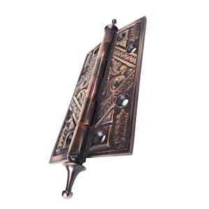 May include: Bronze-colored door hinge with intricate floral and geometric patterns. The hinge has a decorative finial at the top and bottom. The metal hardware is designed for doors and cabinets.