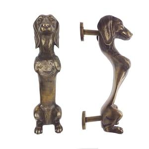 May include: Bronze-colored door handle shaped like a dachshund dog. The handle is detailed with the dog's features, including its face, ears, and body. The handle is mounted on a round plate.