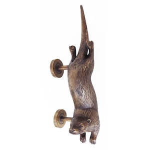 May include: A bronze-colored door knocker in the shape of an otter, hanging upside down. The otter has detailed fur texture and is attached to the door with two circular brass-colored plates. The knocker is a decorative home decor item.