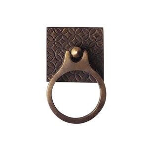May include: A bronze-toned cabinet pull with a square backplate featuring a repeating floral pattern. The pull has a ring-shaped handle and a decorative, curved support. The hardware is designed for furniture and adds a vintage touch.