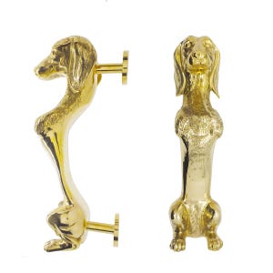 May include: Two gold-colored dachshund door handles. One handle is shown in profile, the other is facing forward. The handles are detailed with the dog's features and have a shiny finish. The handles are mounted on a white background.