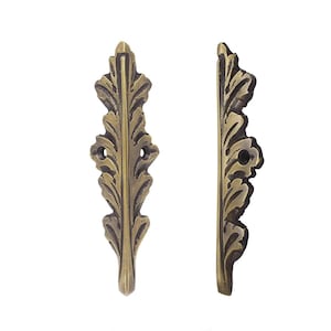May include: Two antique brass-coloured leaf-shaped decorative hardware pieces. Each piece has a detailed, textured design with a series of leaf-like patterns and two small holes for mounting. The hardware is set against a white background.