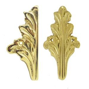 May include: Two ornate, gold-colored leaf-shaped decorative furniture hardware pieces. One is angled, and the other is upright. Each has a detailed, textured surface and small holes for mounting. The hardware is likely for cabinets or other decorative applications.