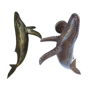 May include: Two bronze whale door knockers. One whale is facing up and the other is facing down. Both whales have detailed textures and are in a jumping pose.
