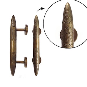 Puede incluir: Tiradores de puerta de color bronce con intrincados patrones grabados. Los tiradores tienen forma de bala con una base redondeada y una punta. El diseño incluye una placa de montaje circular y una barra de conexión. La imagen también incluye un primer plano de los detalles.