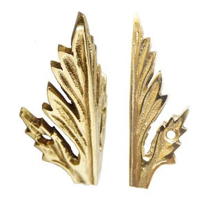 May include: Two gold-toned metal leaf-shaped decorative corner brackets. Each bracket has a detailed, intricate design with a pointed tip and a hole at the base.