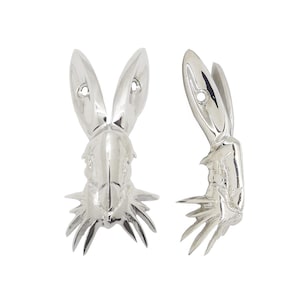 May include: Two silver-colored rabbit head sculptures. One is viewed from the front, the other from the side. Each has long ears with a hole near the top and a detailed face and neck. The sculptures have a shiny, reflective surface and are set against a white background.