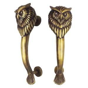 May include: Two antique brass door handles. Each handle features a detailed owl head at the top, with intricate feather detailing. The handles have a curved design and are attached to circular mounting plates. The overall color is a warm, aged brass.
