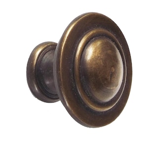May include: A close-up of a bronze-colored cabinet knob. The knob has a rounded, layered design with a central, oval-shaped protrusion. The finish has a slightly aged appearance, suggesting a vintage or antique style. The hardware is likely for furniture.