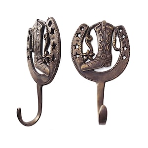 May include: Two bronze-colored metal wall hooks. Each hook features a cowboy boot and star design within a horseshoe shape. The hooks are designed for hanging items and have a rustic, Western theme.