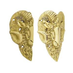 Puede incluir: Dos tiradores de puerta de latón con cabeza de carnero. Cada tirador presenta una cabeza de carnero detallada con cuernos curvos y un asa rectangular. Los tiradores son de color dorado brillante y están montados sobre un fondo blanco.