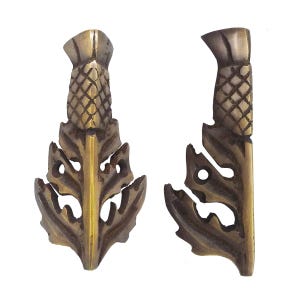 May include: Two antique brass-toned thistle-shaped decorative elements. Each piece features a detailed thistle head and stylized leaf design. The metal has a warm, aged patina, suggesting vintage craftsmanship. The pieces are isolated against a white background.
