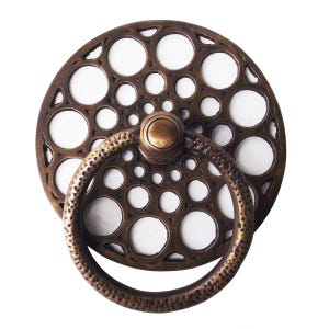 Puede incluir: Tirador de armario en tono bronce con una placa trasera circular con un patrón de agujeros de varios tamaños. Un tirador de anillo a juego está unido al centro, creando una pieza de herraje decorativa y funcional.