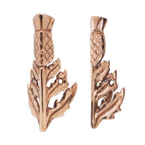 Puede incluir: Dos pendientes en forma de cardo de color bronce. Cada pendiente presenta una cabeza de flor de cardo detallada y hojas intrincadamente diseñadas. Los pendientes se presentan sobre un fondo blanco, resaltando su acabado metálico y su detallada artesanía.