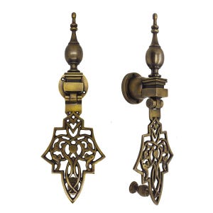 Może przedstawiać: Dwa antyczne mosiężne uchwyty do drzwi z ozdobnymi, ażurowymi wzorami. Każdy uchwyt ma dekoracyjne zwieńczenie, zawiasowy mechanizm i stylizowany, symetryczny wzór. Białe tło.