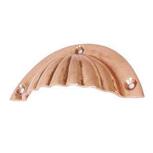 May include: A copper-colored, half-moon shaped cabinet pull. The pull has a scalloped design on the front and three holes for mounting. The hardware is made of metal and has a shiny finish. This decorative pull is suitable for furniture.