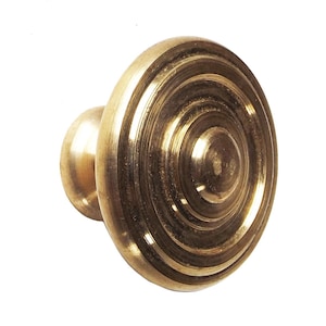 Könnte beinhalten: Ein Messing-Schrankknopf mit kreisförmigem Design. Der Knopf hat einen abgerundeten, zylindrischen Sockel und einen größeren, scheibenförmigen Kopf mit konzentrischen Ringen. Die Oberfläche ist warm golden, vor weißem Hintergrund.