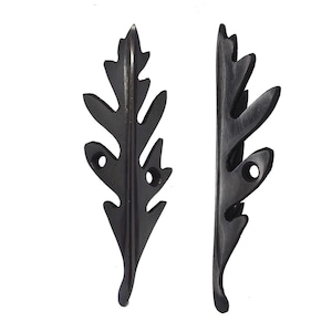 May include: Two dark brown, leaf-shaped decorative hardware pieces. Each piece has a detailed, cutout leaf design with a pointed tip and two small holes. The hardware is likely for furniture or decorative purposes.