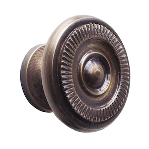 Puede incluir: Un pomo de armario en tono bronce con un diseño circular. El pomo presenta un anillo exterior texturizado y estriado y un centro liso y redondeado. La ferretería tiene un aspecto clásico y antiguo.