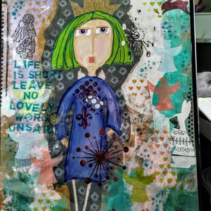 Può includere: Un'opera d'arte a tecnica mista incorniciata raffigurante una figura centrale con capelli verdi e una corona dorata che indossa un abito blu. Lo sfondo include sagome di conigli, angeli e cuori. Il testo a sinistra recita: LIFE IS SHORT LEAVE NO LOVELY WORD UNSAID.