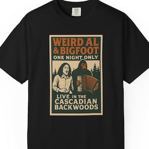 May include: Black t-shirt with a vintage-style graphic. The design features the text "WEIRD AL & BIGFOOT" above an illustration of two figures, one playing an accordion. The text "LIVE IN THE CASCADIAN BACKWOODS" is below the image.