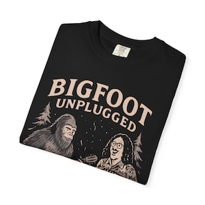 May include: Black t-shirt featuring a Bigfoot illustration with the text "BIGFOOT UNPLUGGED". The design includes a cartoon Bigfoot and a person playing a guitar. The shirt is made of a soft material.