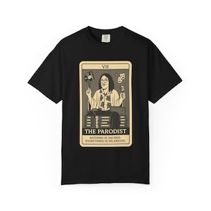 May include: Black t-shirt featuring a tarot card design with the text "THE PARODIST." The card depicts a person holding a microphone and a record, with the words "NOTHING IS SACRED. EVERYTHING IS HILARIOUS."
