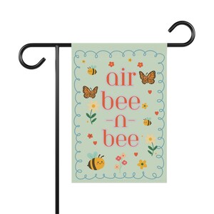 Può includere: Una bandiera da giardino con le parole "air bee -n- bee" in rosso, con illustrazioni di api, farfalle, fiori e cuori su sfondo verde chiaro. La bandiera è sostenuta da un supporto in metallo nero.
