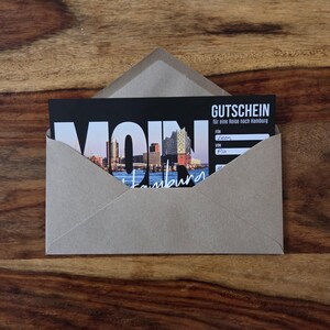 K&ouml;nnte beinhalten: Ein brauner Kraftpapierumschlag mit einem schwarzen Gutschein f&uuml;r eine Reise nach Hamburg. Der Gutschein zeigt ein Foto der Hamburger Skyline, das Wort MOIN in gro&szlig;en wei&szlig;en Buchstaben und den Text GUTSCHEIN f&uuml;r eine Reise nach Hamburg.