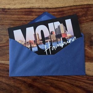 K&ouml;nnte beinhalten: Eine rechteckige Postkarte mit einem Foto der Hamburger Skyline, auf dem der gro&szlig;e wei&szlig;e Schriftzug MOIN und der kursive wei&szlig;e Schriftzug Hamburg zu sehen sind, in einem blauen Umschlag mit einer nach oben zeigenden dreieckigen Klappe.