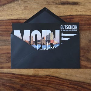 K&ouml;nnte beinhalten: Ein schwarzer Umschlag mit einem Gutschein f&uuml;r eine Reise nach Hamburg. Der Gutschein zeigt ein Foto der Hamburger Skyline, das Wort MOIN in gro&szlig;en wei&szlig;en Buchstaben und den Text GUTSCHEIN f&uuml;r eine Reise nach Hamburg mit handschriftlichen Namen.