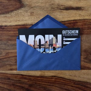 K&ouml;nnte beinhalten: Ein blauer Umschlag mit einem schwarzen Gutschein f&uuml;r eine Reise nach Hamburg. Der Gutschein zeigt ein Foto der Hamburger Skyline, den Text GUTSCHEIN, MOIN und Hamburg sowie die handschriftlich eingetragenen Namen Leon und Pia.