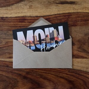 K&ouml;nnte beinhalten: Ein hellbrauner Papierumschlag mit einer Postkarte, die das Wort MOIN in gro&szlig;en wei&szlig;en Buchstaben zeigt, sowie ein fotografisches Stadtbild von Hamburg mit der Elbphilharmonie und dem Schriftzug Hamburg in wei&szlig;er Schreibschrift.