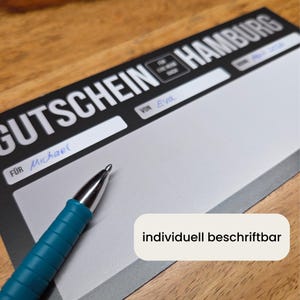 K&ouml;nnte beinhalten: Ein rechteckiger Gutschein mit schwarzer Kopfzeile und dem wei&szlig;en Text GUTSCHEIN HAMBURG. Das Formular enth&auml;lt Felder mit der Beschriftung F&Uuml;R, VON und DATUM, ausgef&uuml;llt mit den Namen Michael und Eva in blauer Tinte. Ein t&uuml;rkisfarbener Kugelschreiber liegt darauf.