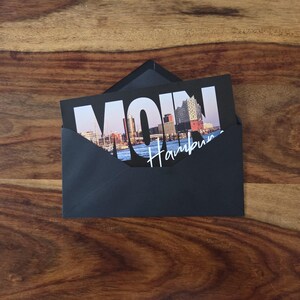 K&ouml;nnte beinhalten: Ein dunkelgrauer Umschlag mit einer Postkarte, die eine Fotografie der Hamburger Skyline bei Sonnenuntergang zeigt. Das Wort MOIN ist in gro&szlig;en wei&szlig;en Buchstaben &uuml;ber das Stadtbild gedruckt, darunter steht Hamburg in wei&szlig;er Schreibschrift.